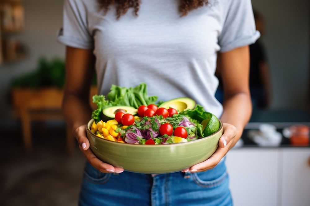 Woman in t-shirt and jeans standing and holding vegan bowl or salad bowl in kitchen adult food midsection. AI generated…