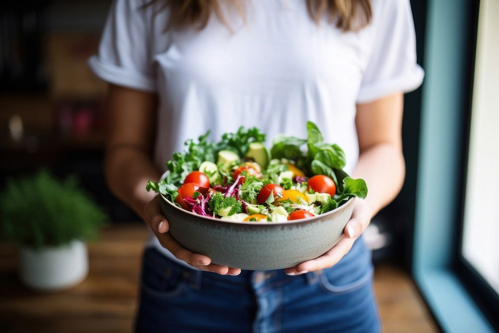 Woman in t-shirt and jeans standing and holding vegan bowl or salad bowl in kitchen adult plant food. AI generated Image by…