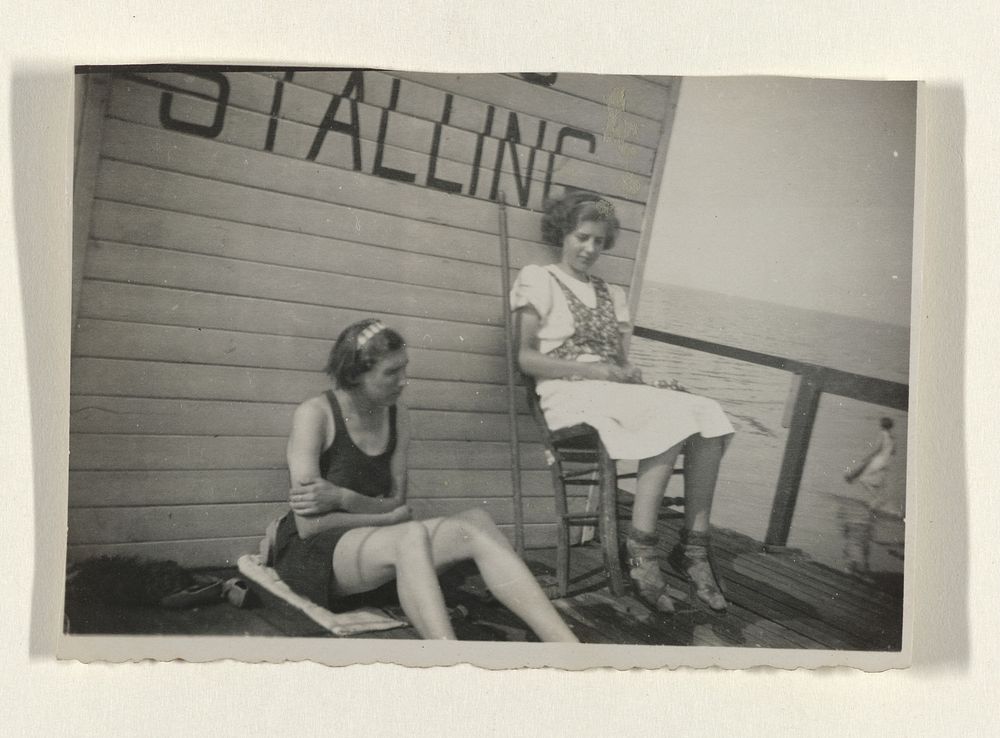 Twee meisjes zittend tegen de muur van een stalling op een pier in de Zuiderzee (c. 1925 - c. 1950) by anonymous