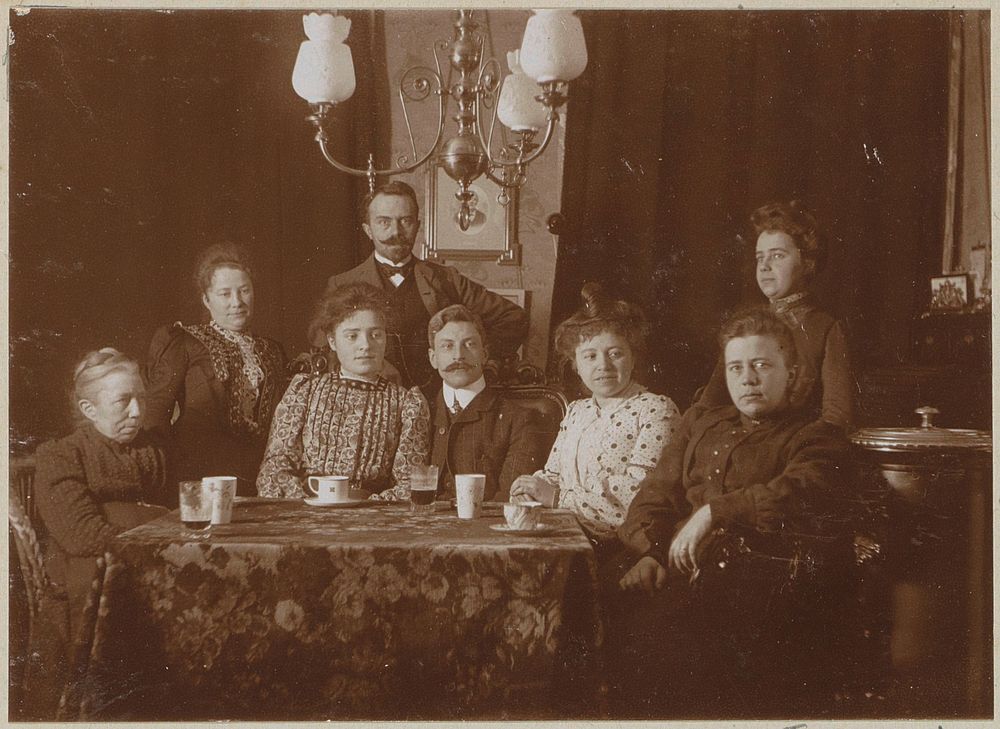 Mannen en vrouwen achter een tafel in een woonkamer (c. 1900 - c. 1910) by anonymous