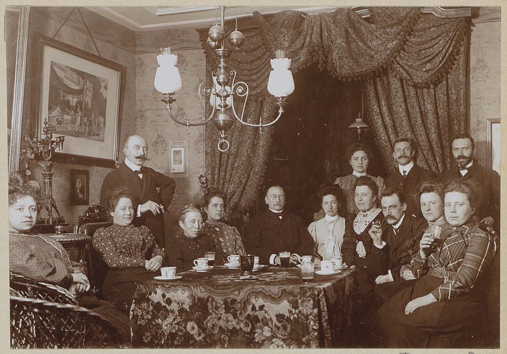 Mannen en vrouwen achter een tafel in een woonkamer (c. 1900 - c. 1910) by anonymous