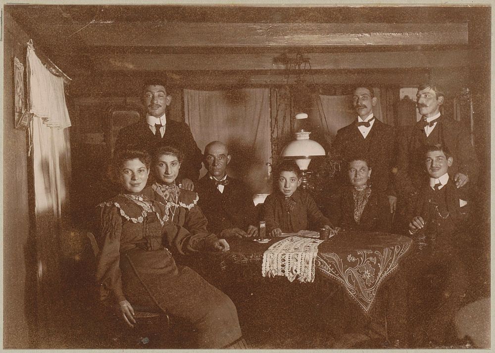 Mannen en vrouwen achter een tafel in een vertrek met balkenplafond (c. 1900 - c. 1910) by anonymous