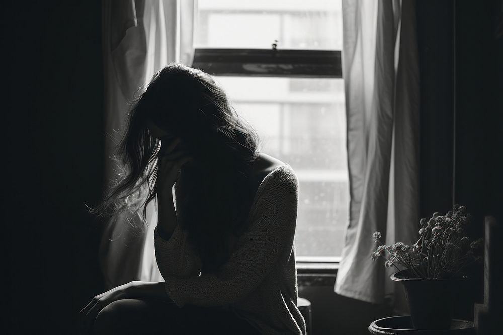 A girl sitting in the room covering her face window worried black.