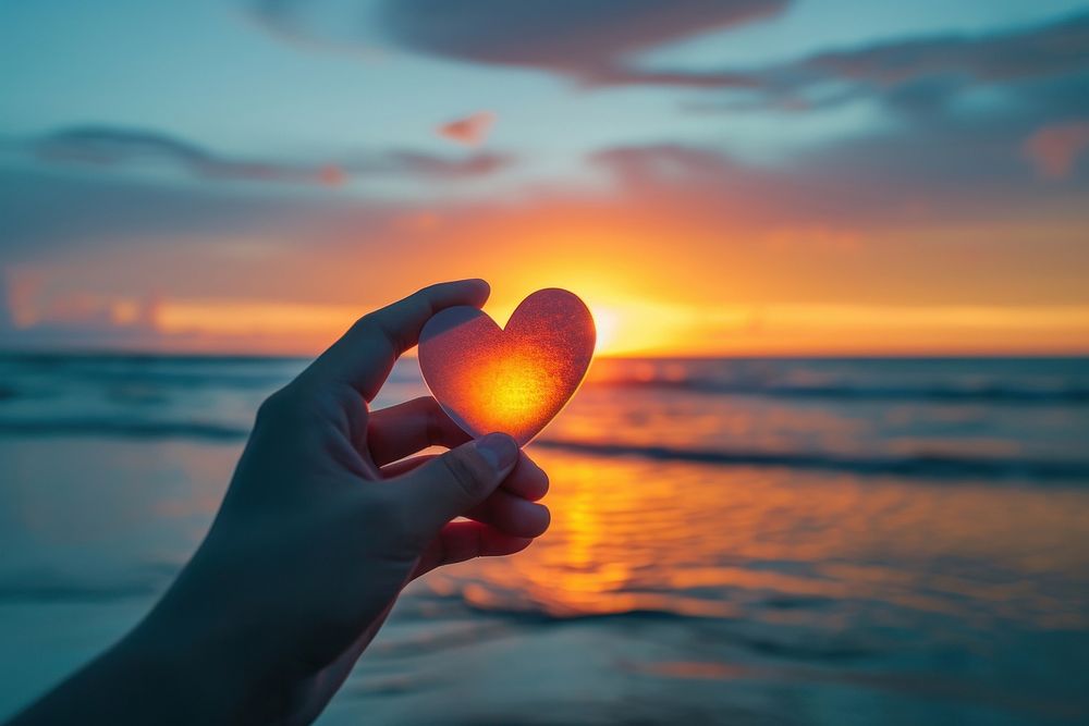 Hand holding a cut out of heart shaped paper against a sunset beach sunlight sky tranquility.