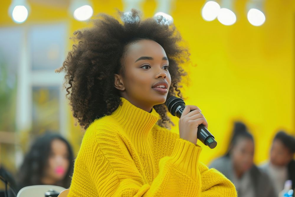 An african american woman wearing yellow sweater holding up microphone speaking in a conference adult women hairstyle.