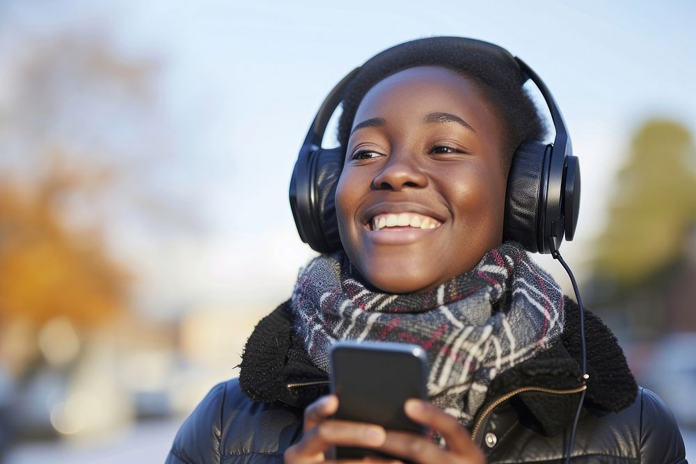 Happy young black woman holding mobile phone enjoying music listening through wireless headphones on footpath headset smile…