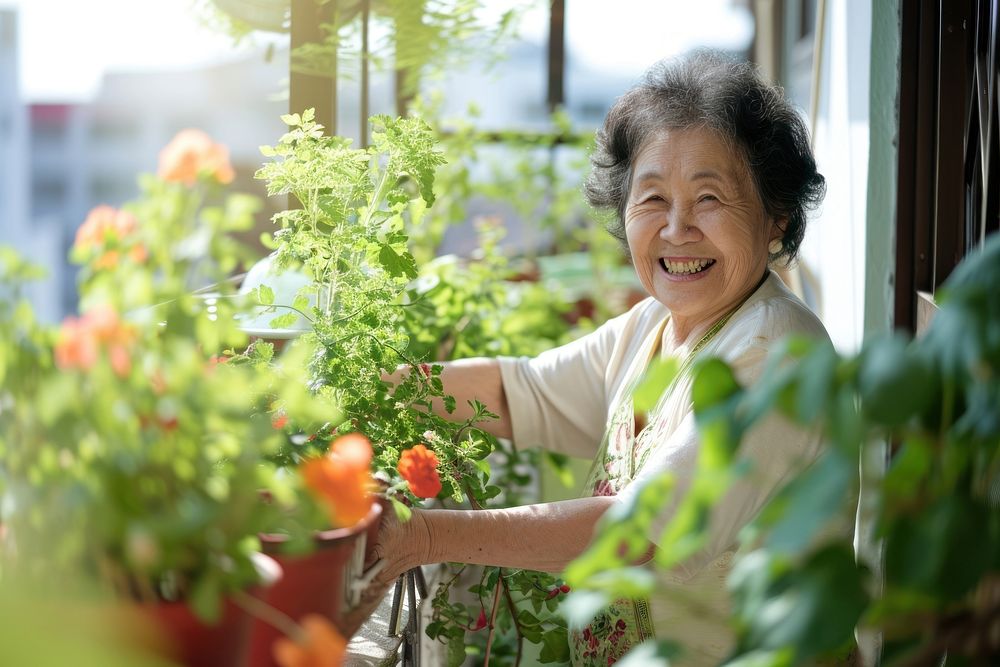 Asian older woman smiling warmly while tending to her potted plants on a sunny balcony gardening cheerful outdoors. AI…