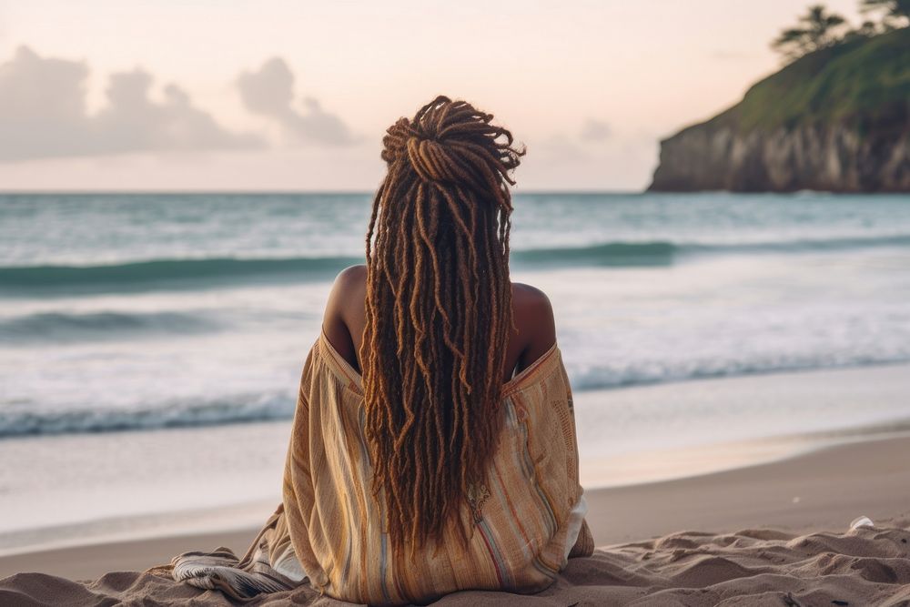 An African american woman with dreadlock hair sitting relaxed on a beach looking at the sea outdoors nature summer.