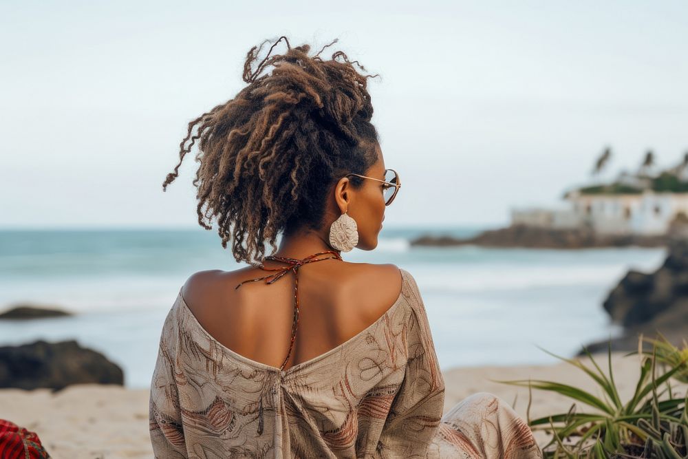 An African american woman with dreadlock hair sitting relaxed on a beach looking at the sea summer adult back.