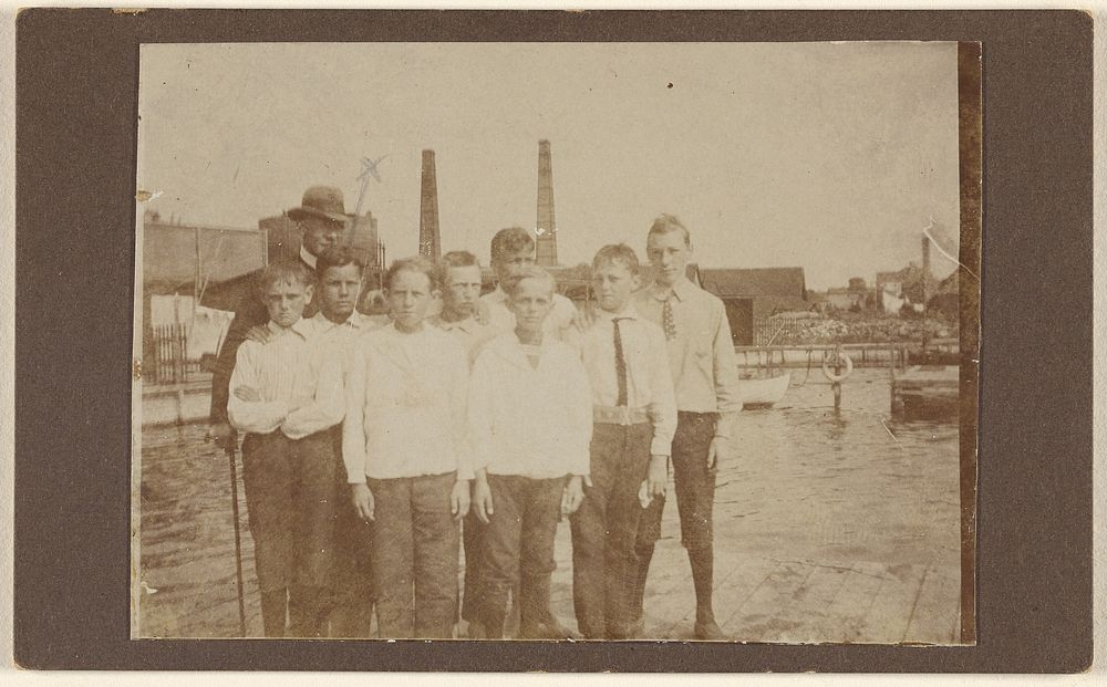 Eight young men standing on a dock near a river, with a man wearing a hat