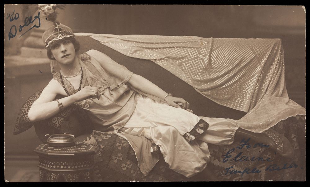A man in drag poses wearing delicate attire; curled up on a piece of furniture. Photographic postcard, 191-.