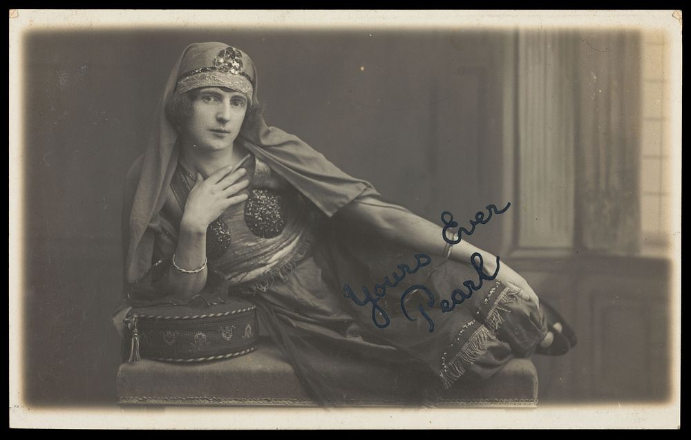 A man in drag, poses wearing delicate attire; curled up on a piece of furniture. Photographic postcard, 191-.