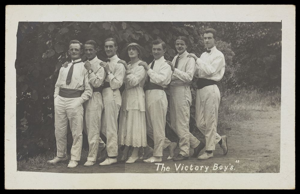 Entertainers, one in drag, pose in a line in front of foliage. Photographic postcard, 191-.