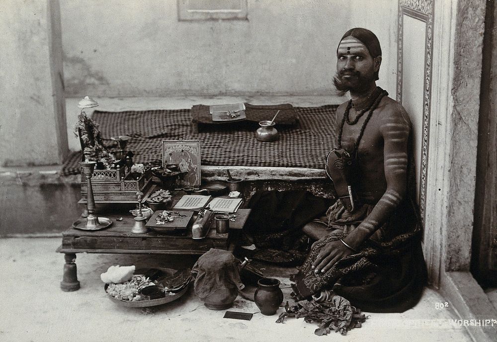 An Indian man wearing elaborate jewellery and make-up, squatting in front of a small table on which is a shrine, perhaps in…