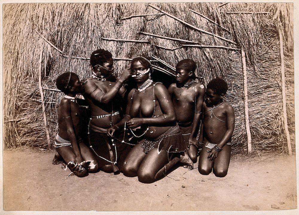 Africa: African women adorning themselves with beads, outside a kraal hut. Albumen print.
