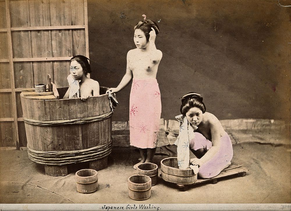 Three Japanese women washing: One in a wooden tub, one standing and one crouching over a small wooden tub on the floor.…