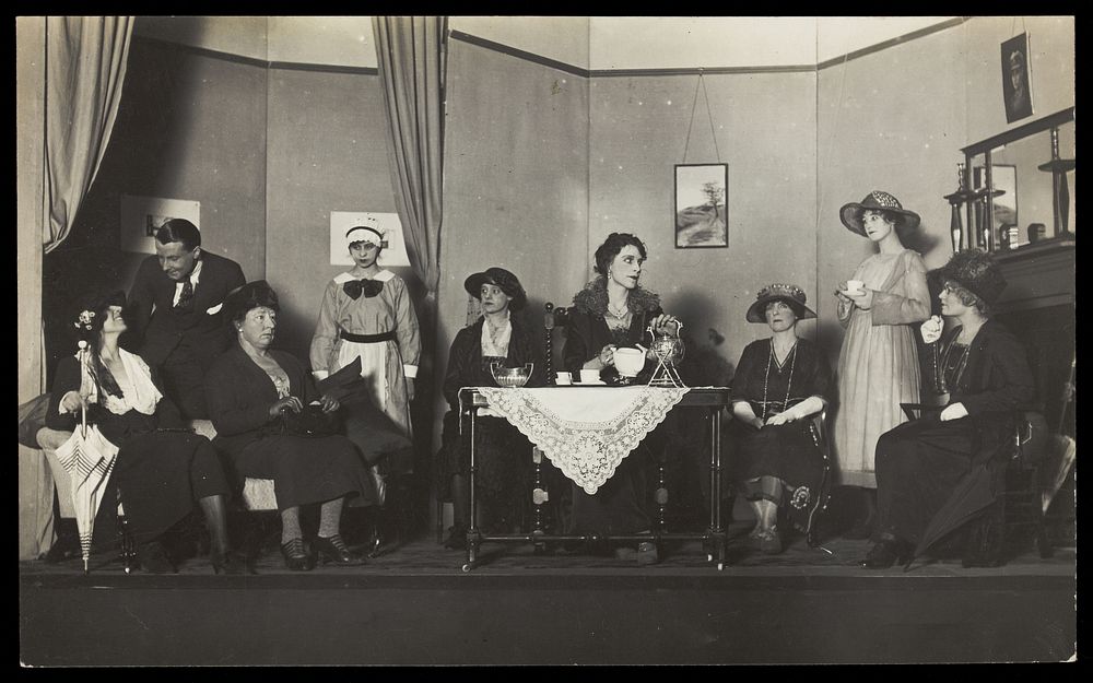 Amateur actors, some in drag, are having tea on stage. Photographic postcard, 192-.