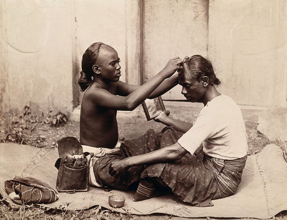 Two Indian men sitting cross-legged; one appears to be a barber and is shaving the front of the other's head, presumably to…