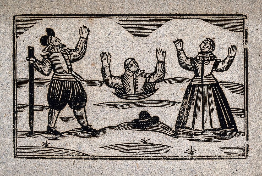A man buried up to his waist holds his hands up above his head: a man and a woman stand on either side watching him in…