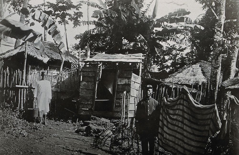 Sierra Leone: wooden cesspool huts, with a uniformed man and a man holding a bucket shown standing in the foreground.…