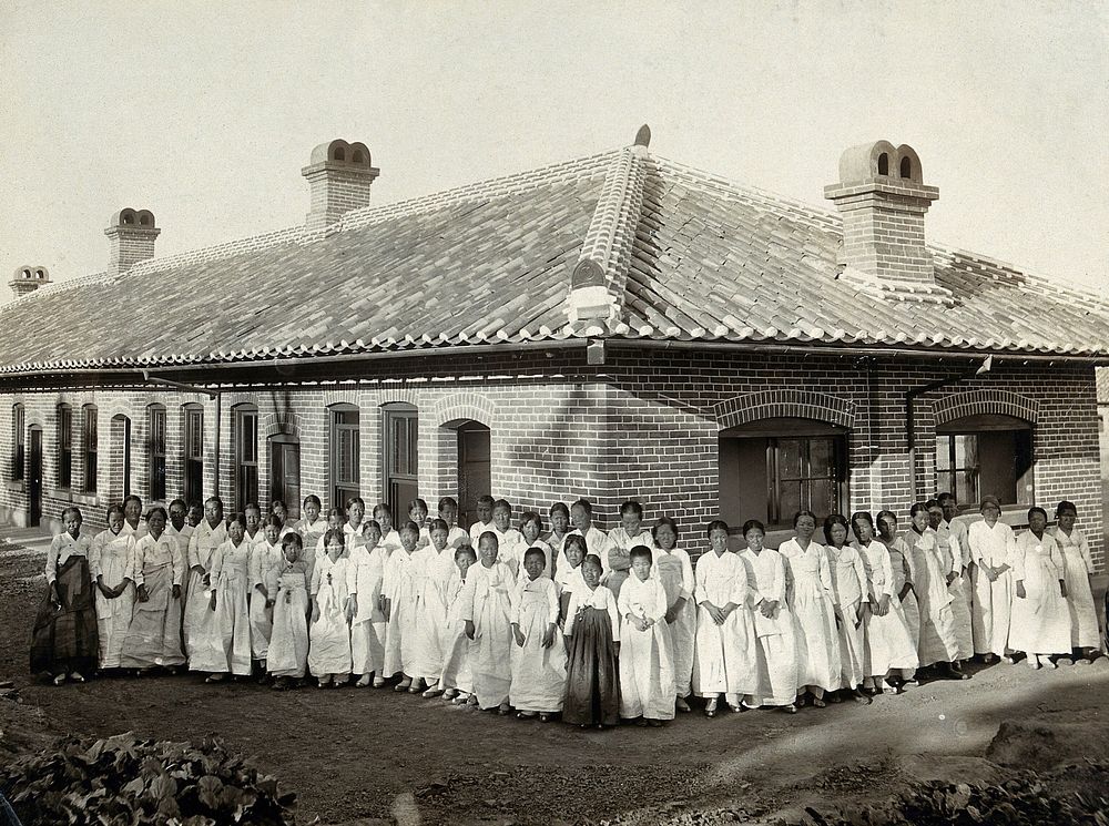 Leper asylum, Taiku, Korea: female patients wearing white robes stand in a group outside a brick building. Photograph…
