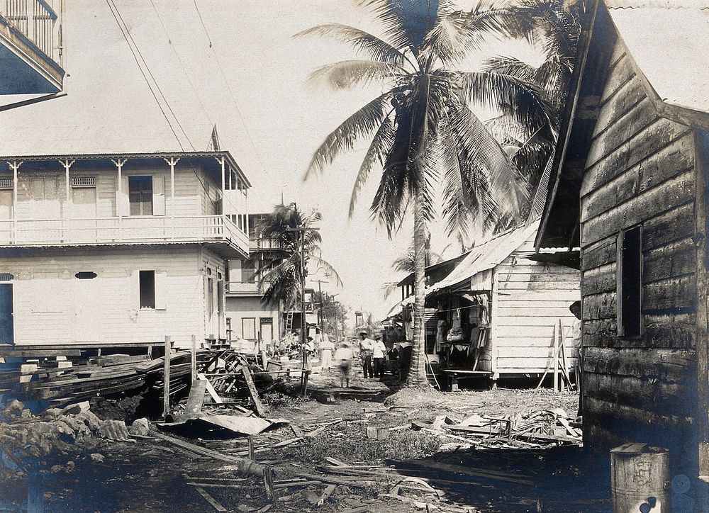 Colón, Panama: unpaved street with wooden houses and palm trees: building debris scattered in foregound; people visible in…