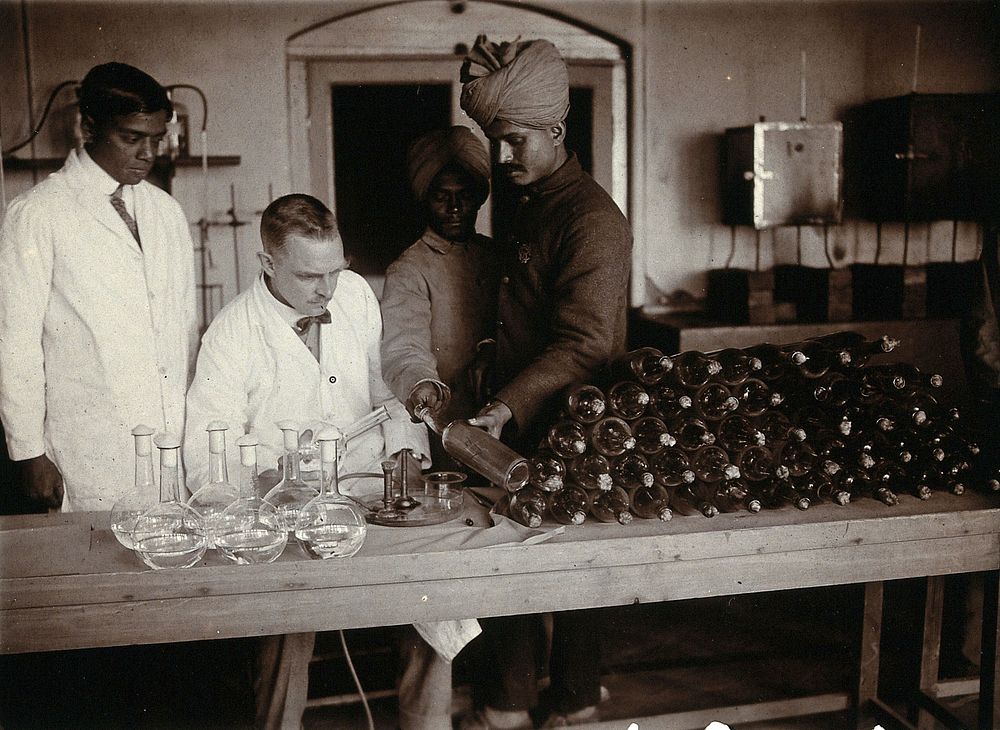 A laboratory with staff (Indian and British ) at work at a table piled high with bottles. Photograph, 1900/1920 .