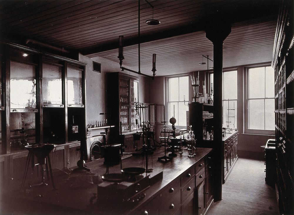 The interior of a pharmaceutical or chemical laboratory, with two long benches in the centre, glass-fronted cabinets and a…