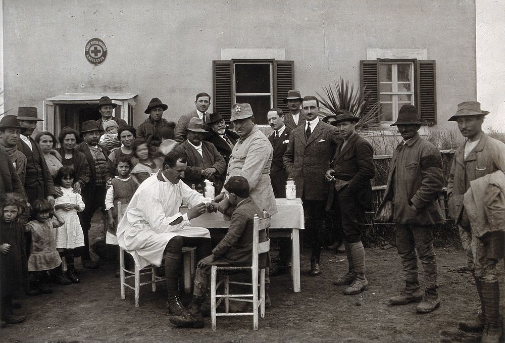 Nettuno , Italy: a medical man taking a blood sample from a boy seated at a small table, surrounded by a crowd of onlookers…