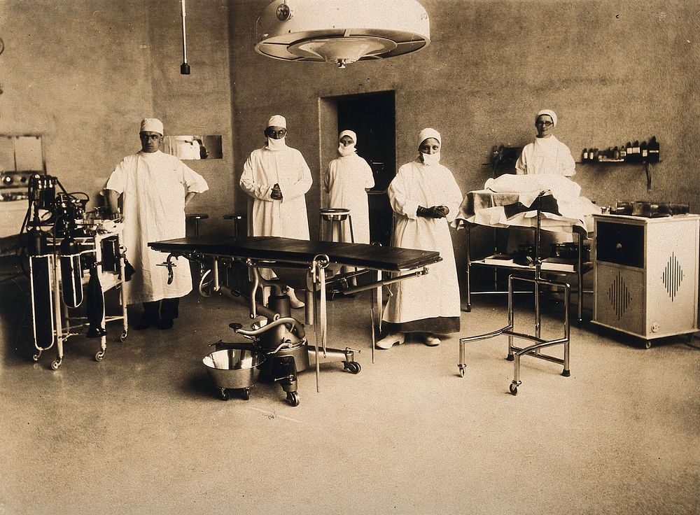 Surgical staff standing in an operating theatre with a lot of stainless steel equipment. Photograph by Bruce & Webster.