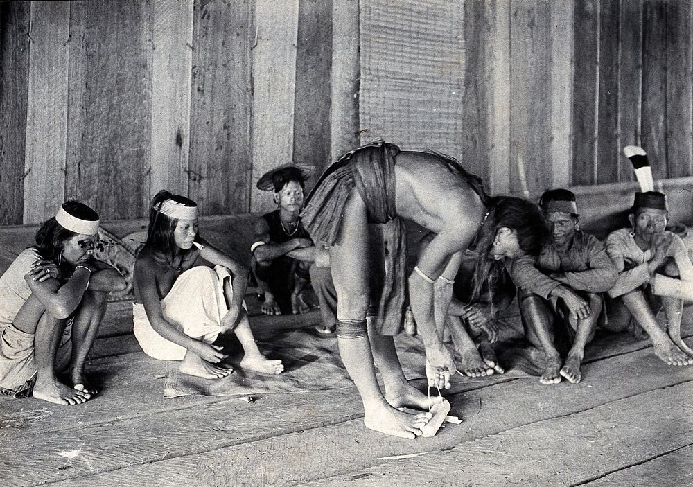 Sarawak: a Kayan man making fire by rubbing cane on wood. Photograph.