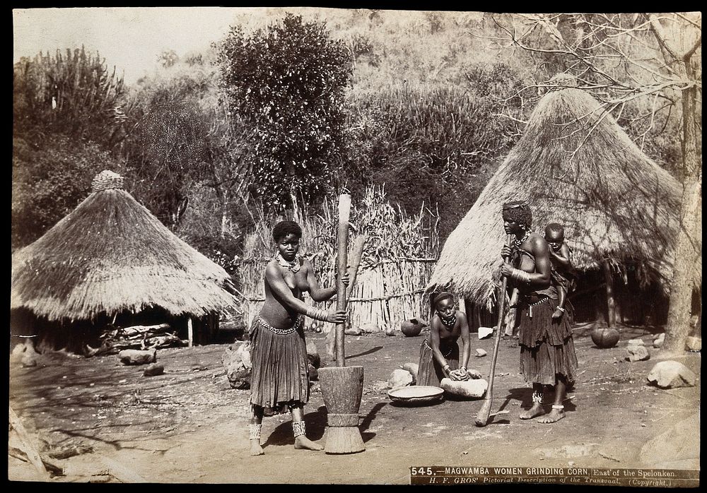 South Africa: Magwamba women grinding corn outside mud huts; one woman works with a baby in a fabric sling on her back.…