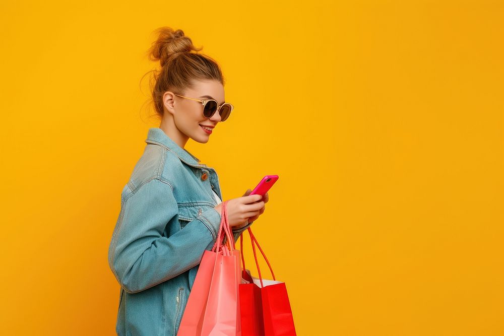 Smiling young lady wear sunglasses using her smartphone while shopping bag handbag red.