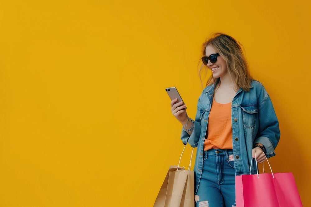 Smiling young lady wear sunglasses using her smartphone while shopping bag shopping bag consumerism.