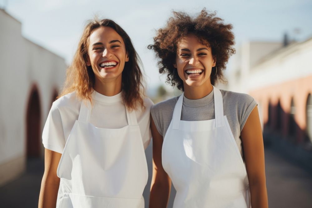 2 women wearing white fabric apron laughing outdoors smile.