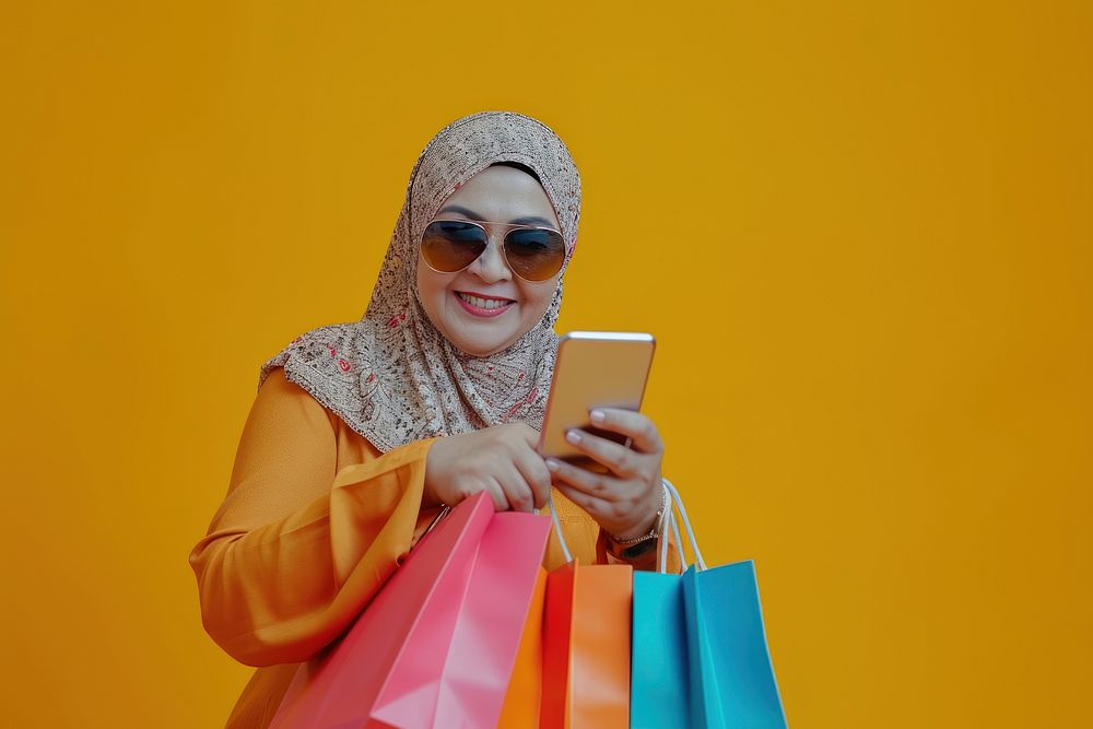 Smiling muslim mature woman wear sunglasses using her smartphone adult shopping bag portability.