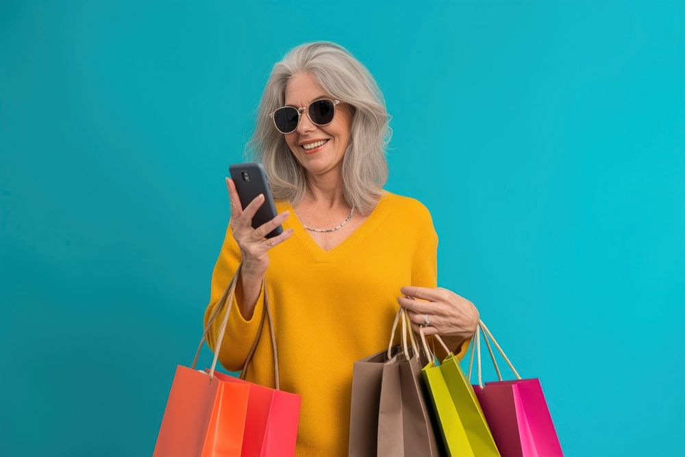 Smiling mature woman wear sunglasses using her smartphone shopping bag handbag.