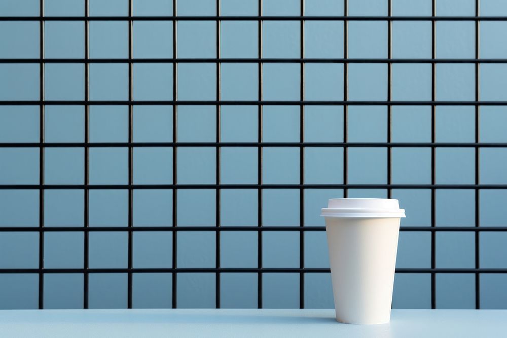 A paper coffee cup is on a black grid fence wall tile blue.