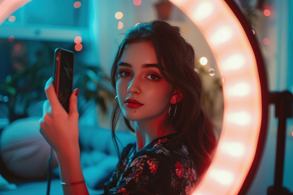 Young woman recording herself taking portrait by selfie ring with smart phone adult photographing illuminated.