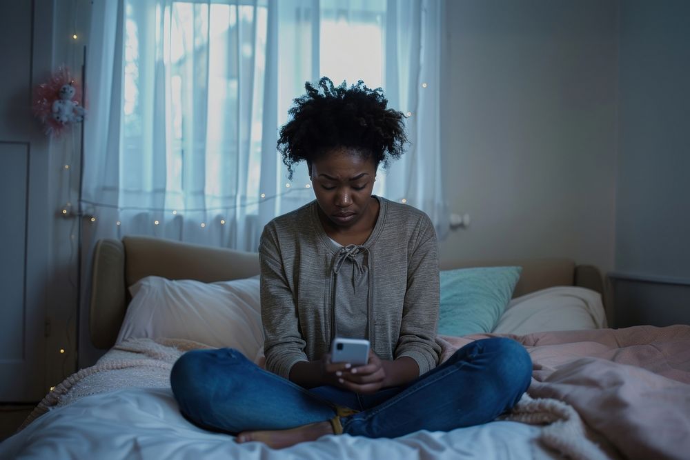 Sad black teenager holding a mobile phone bed electronics furniture.