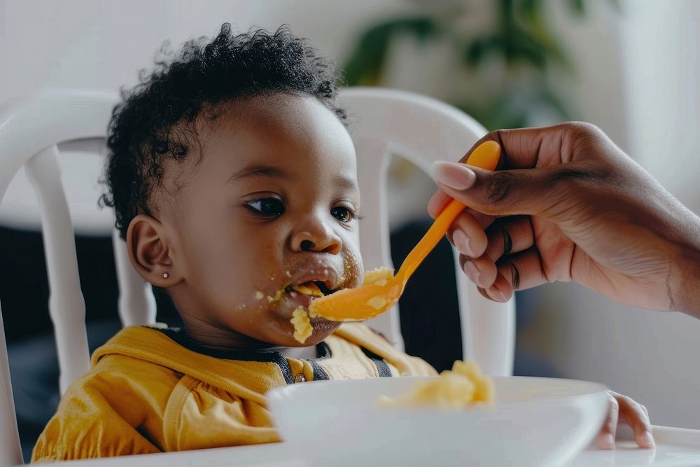 African American toddler sitting in a high chair food cutlery biting.