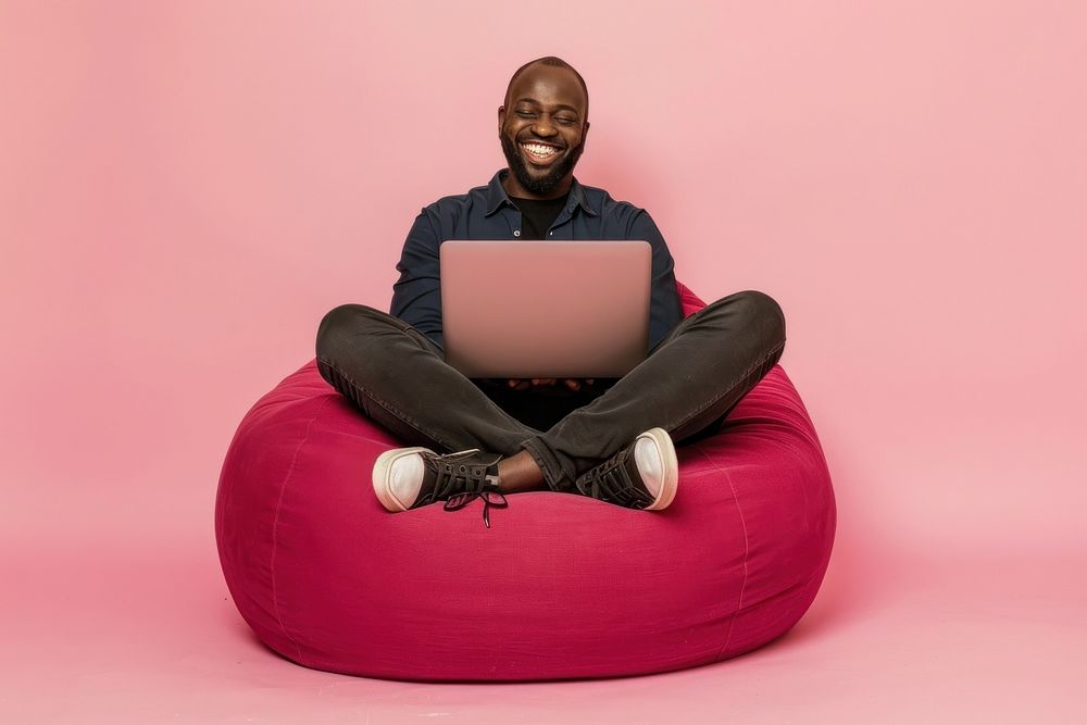 Cheerful black man working on laptop seated on a minimal luxury color beanbag furniture computer sitting.