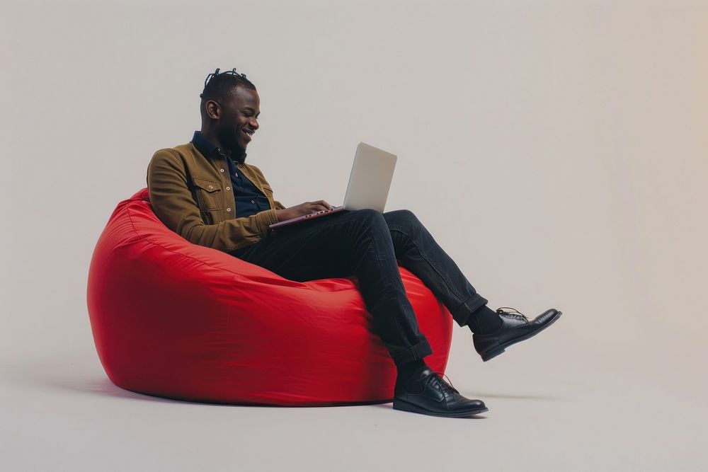 Cheerful black man working on laptop seated on a minimal luxury color beanbag furniture computer adult.