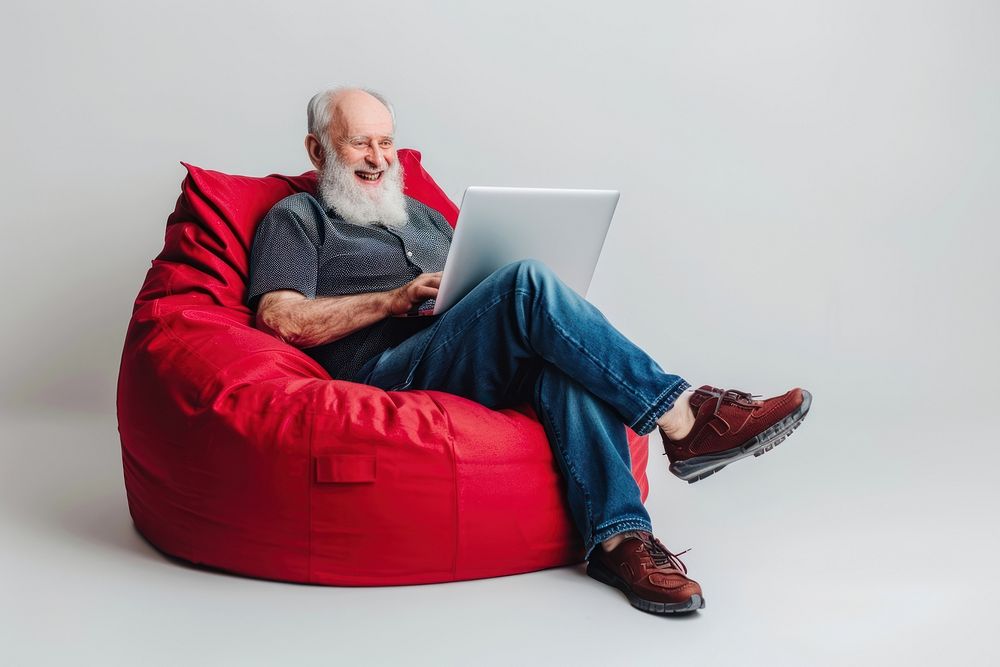 Cheerful old man working on laptop seated on a minimal luxury color beanbag furniture computer adult.