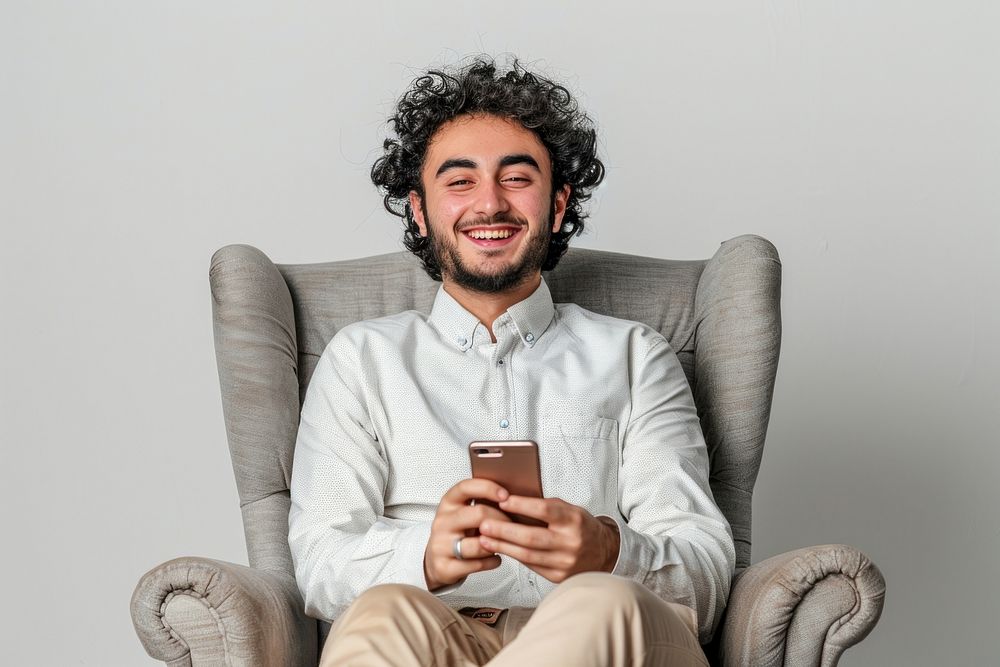 Joyful young male Middle Eastern sitting in armchair with smartphone person human adult.