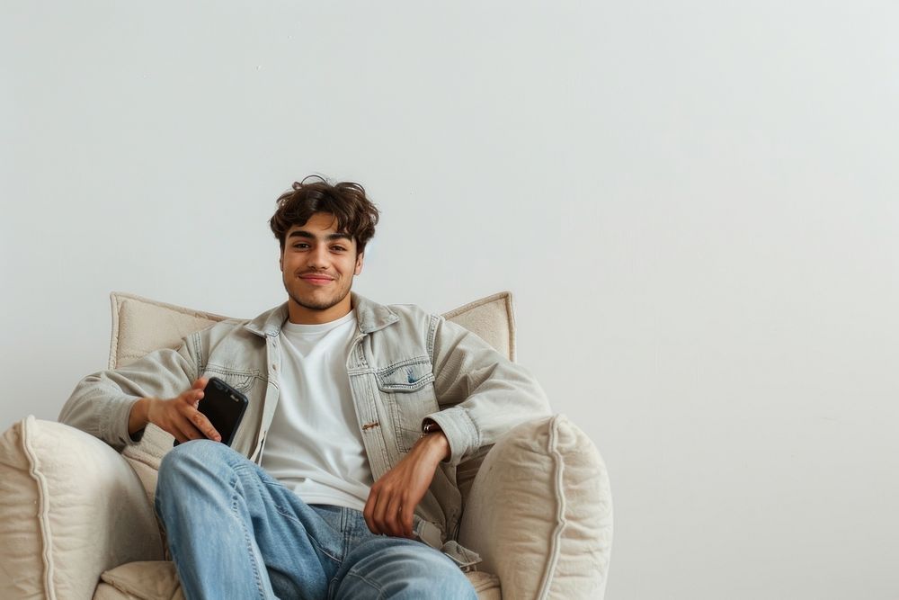 Joyful young male Middle Eastern sitting in armchair with smartphone photo electronics photography.