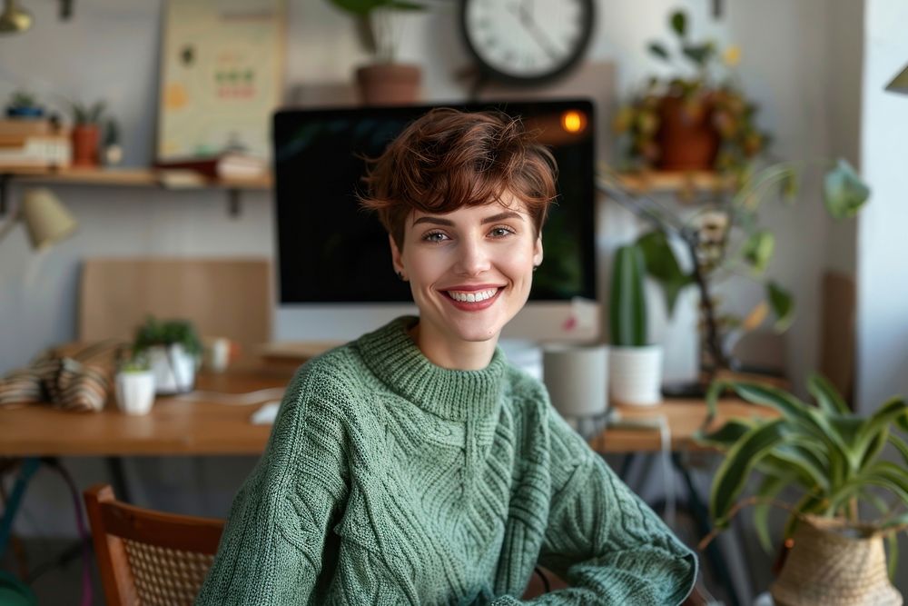 Short hair woman in green mint weater smiling at camera sitting at workstation electronics furniture clothing.