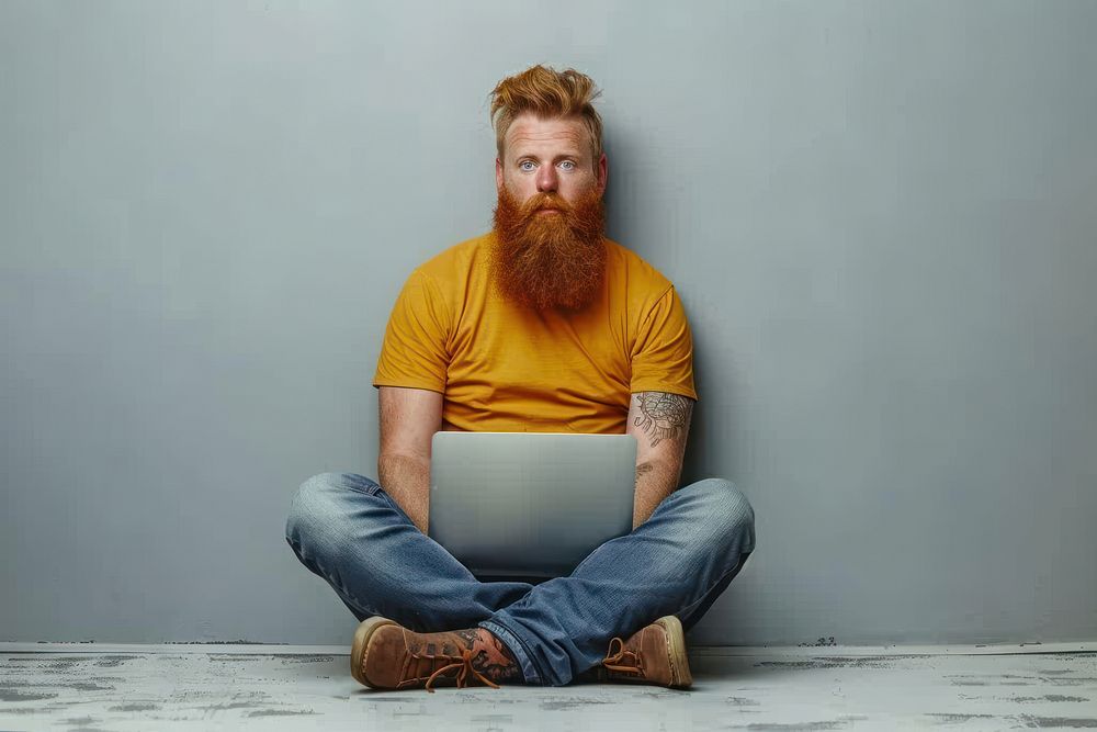 Full body young redhead bearded man in t-shirt casual clothes sitting hold use work on laptop point aside clothing footwear…