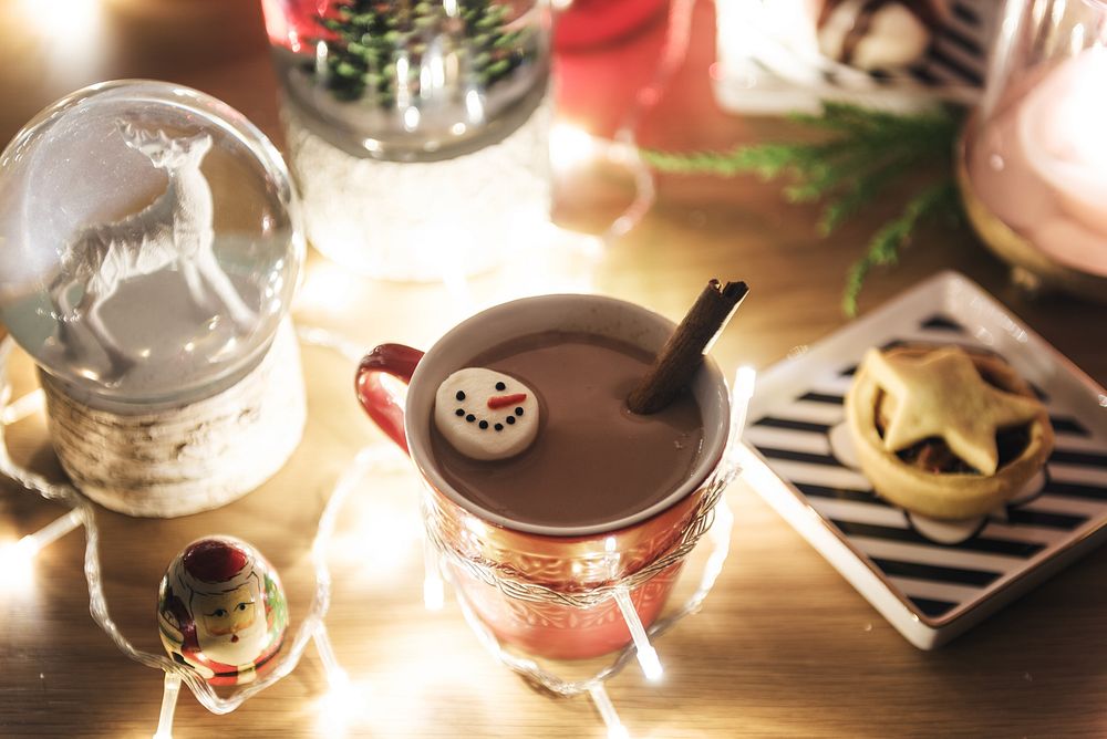 Festive hot chocolate in a red mug with a snowman marshmallow, surrounded by warm lights. Cozy holiday drink, perfect for…