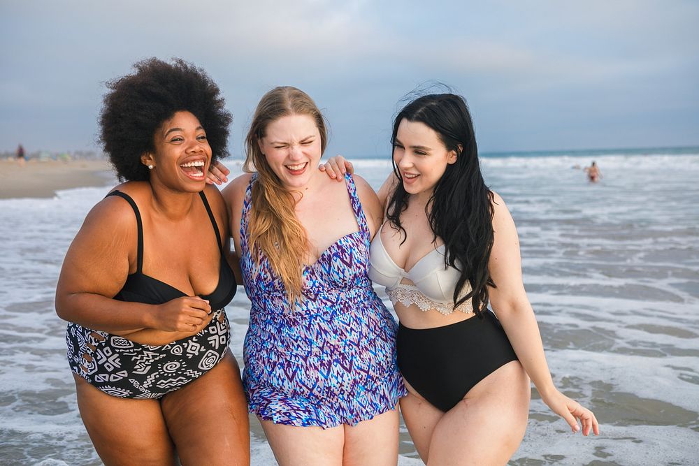 Diverse three women enjoying a day at the beach, laughing and walking by the ocean. Women friends wear swimsuits and appear…